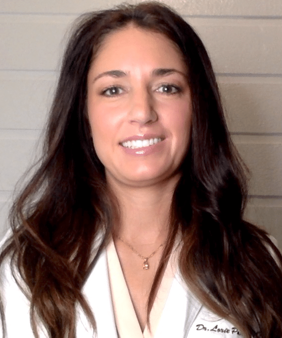 A woman with long brown hair wearing a white coat and a light-coloured blouse smiles in front of a light grey wall. As telemedicine is growing in popularity, her embroidered name highlights her expertise in virtual care.
