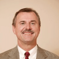 A middle-aged man with short brown hair and a moustache, wearing a brown checked suit, white shirt, and red tie, smiles in front of a plain beige background—far from modelling Must-Have Maternity Dresses for the Summer in 2022.