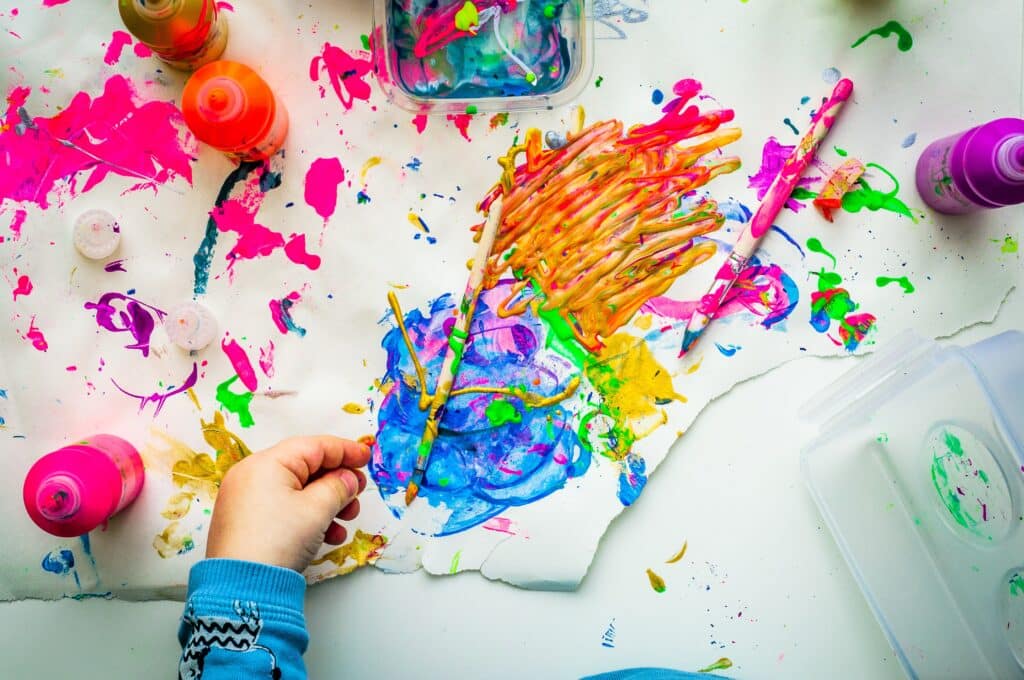 A child’s hand holding a paintbrush, painting colourful abstract shapes on paper—a perfect example of how to boost your child's imagination. Paint bottles and scattered art supplies fill the vibrant, messy workspace.