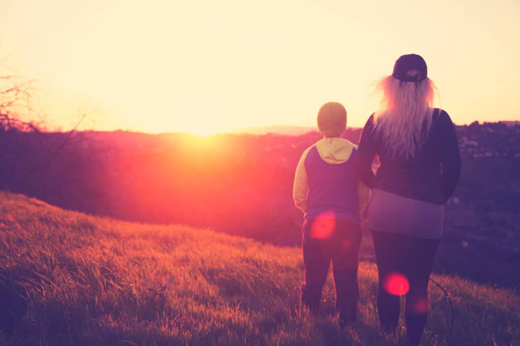 Two people, one adult and one child, stand side by side on a grassy hill at sunset, facing the glowing horizon with warm sunlight casting a soft, golden glow over the landscape.