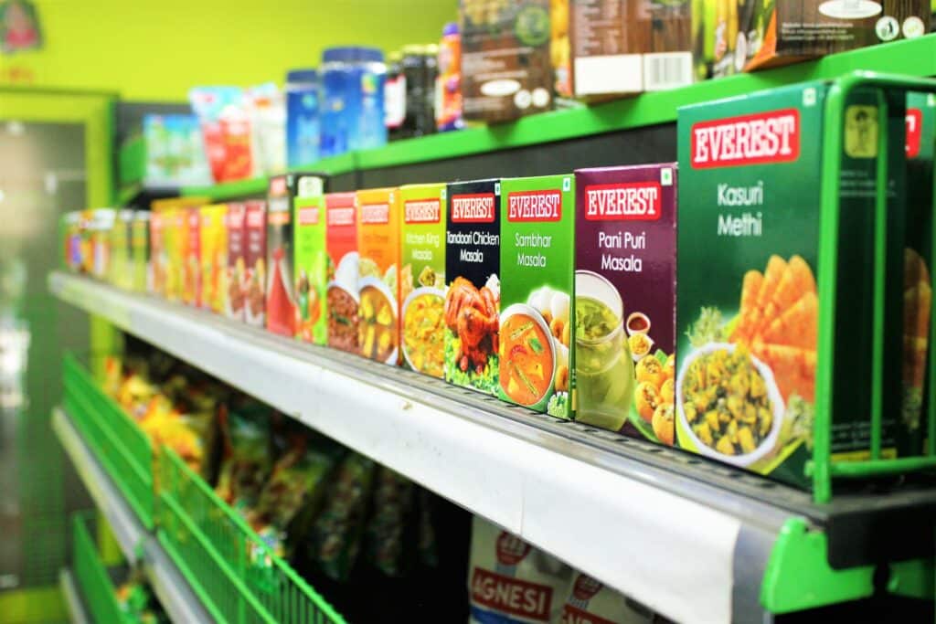 Boxes of Everest brand Indian spices and masalas are neatly arranged on a green shelf in a grocery shop, reflecting excellent customer service with clearly displayed, colourful packaging and other products in the background.
