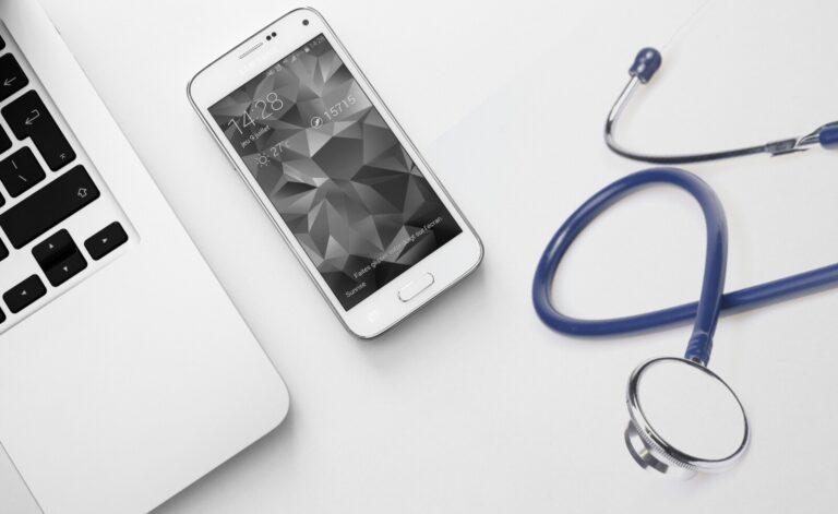 A white smartphone, a silver laptop, and a blue stethoscope are placed on a white surface, suggesting a connection between technology and healthcare.