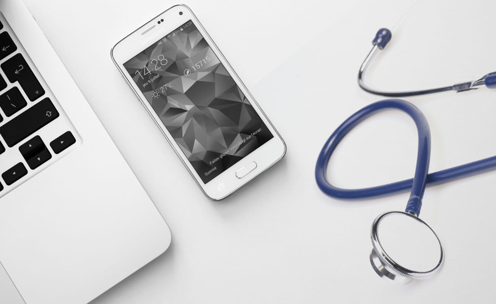 A white smartphone, a silver laptop, and a blue stethoscope are placed on a white surface, suggesting a connection between technology and healthcare.