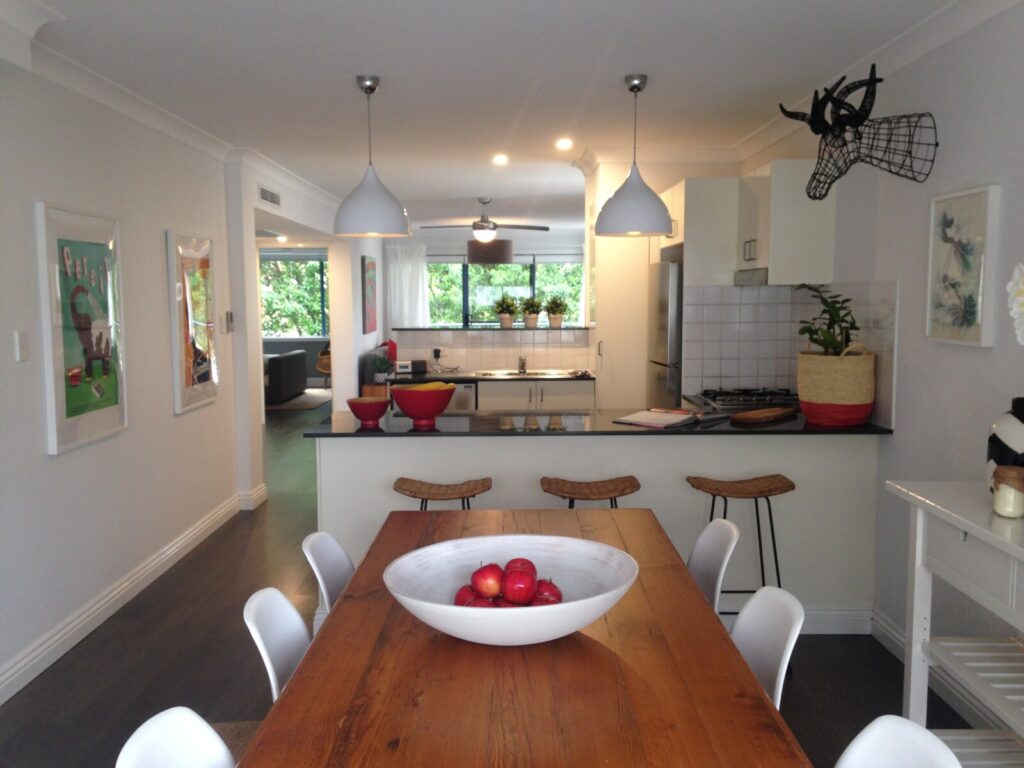 A modern kitchen and dining area with a wooden table, white chairs, a bowl of apples, bar stools, pendant lights, and decorative art on the walls. The kitchen is bright with plants on the windowsill.