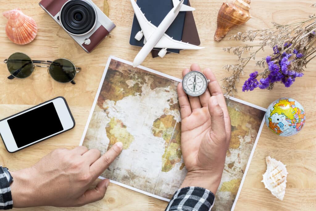 A person points to a world map with one hand and holds a compass in the other. Nearby are a mobile phone, sunglasses, camera, globe, passport, toy aeroplane, seashells, and purple flowers on a wooden table.