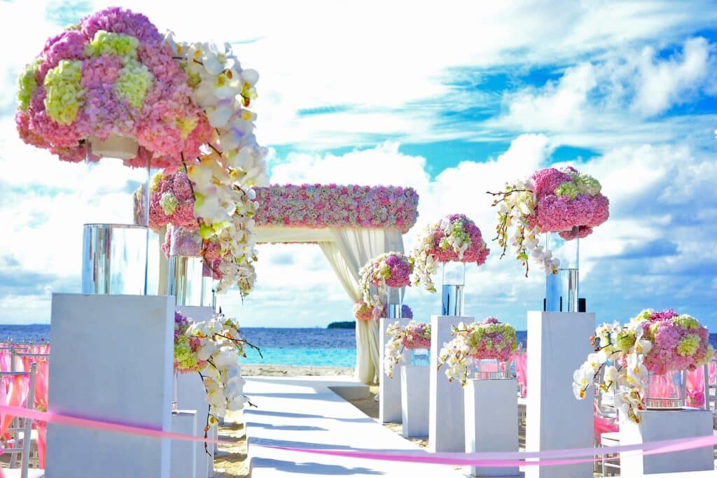 A beach wedding set-up with white pillars topped with pink and white flower arrangements, a flower-adorned arch, and a clear blue sky shows what it takes to have a truly “You-Nique” wedding: personalised touches in every stunning detail.