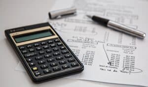 A black calculator sits on a document filled with numbers and handwritten notes, next to an uncapped silver pen. The paper's tables and circled figures suggest it's time to Get Into The Saving Money Mindset through careful financial planning.