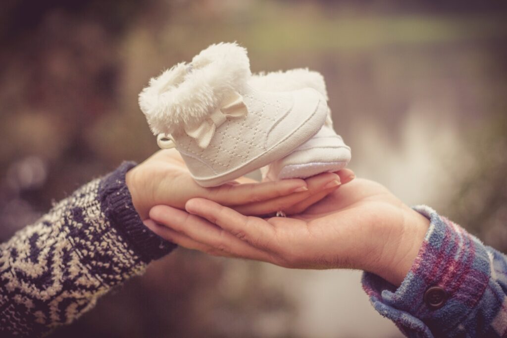 Two hands hold a pair of small white baby shoes with fur trim and bows, symbolising an upcoming arrival. The background is blurred, drawing focus to the shoes.
