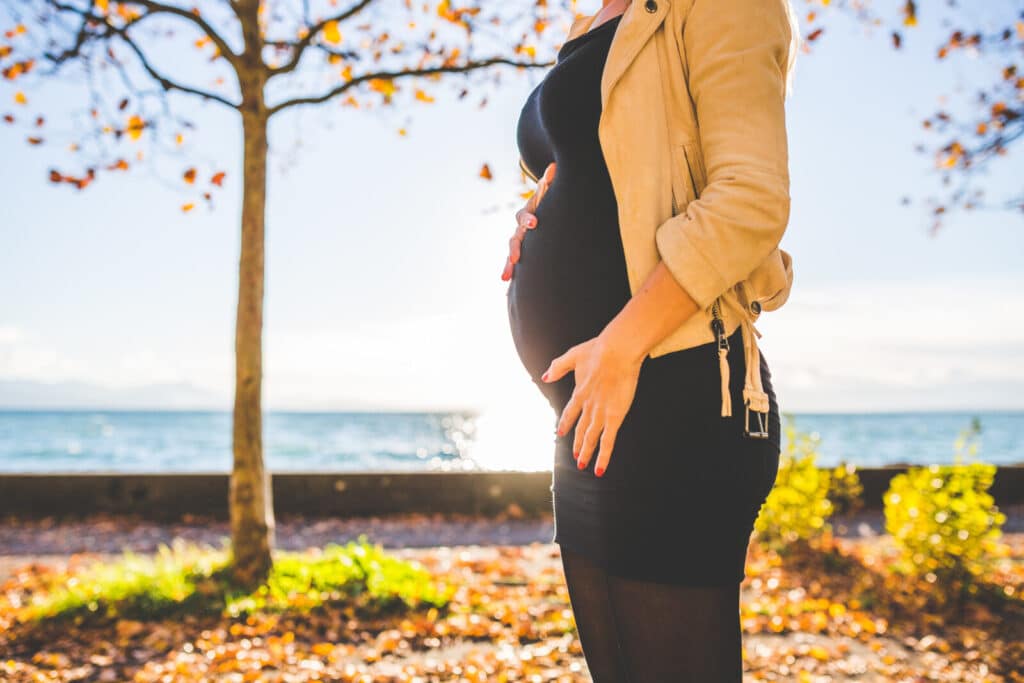 A pregnant woman in a black dress and tan jacket stands outdoors, gently holding her bump. Autumn leaves cover the ground, and a tree and body of water are visible in the background.