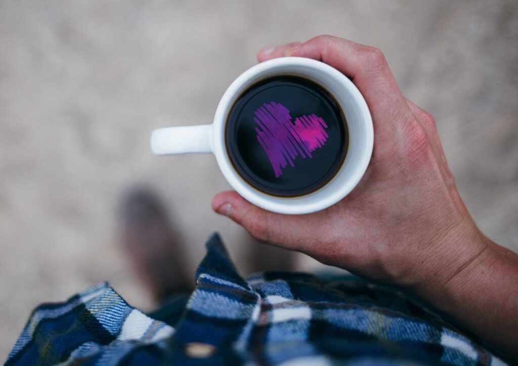 A person holding a white mug of coffee viewed from above, with a pink heart shape reflected in the surface of the coffee. The person is wearing a blue checked shirt.