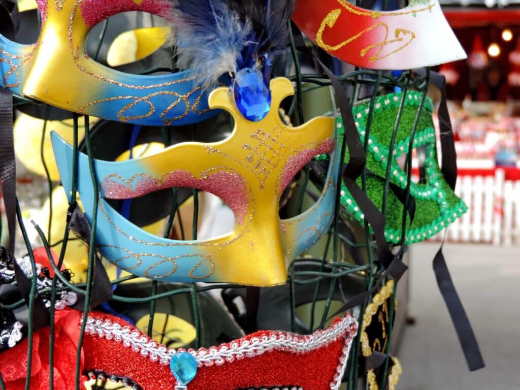 Colourful masquerade masks, including gold, blue, and green designs with glitter, feathers, and gemstones, are displayed on a stand at an outdoor market with white picket fences in the blurred background.