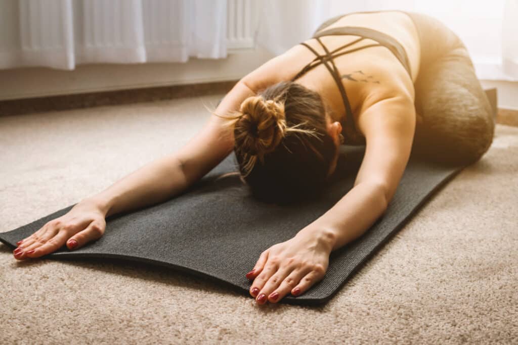A woman practises yoga indoors, stretching forward on a black mat with her arms extended and forehead touching the mat, sunlight streaming in from a nearby window.