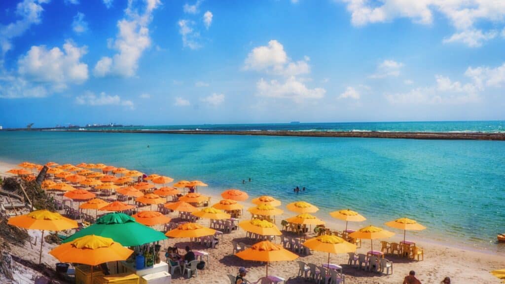 Crowded beach with many yellow and a few green umbrellas and plastic tables and chairs. People swim in the clear turquoise water under a bright blue sky with scattered clouds—take note of Essential Travel Tips for 2022 to make the most of your visit.
