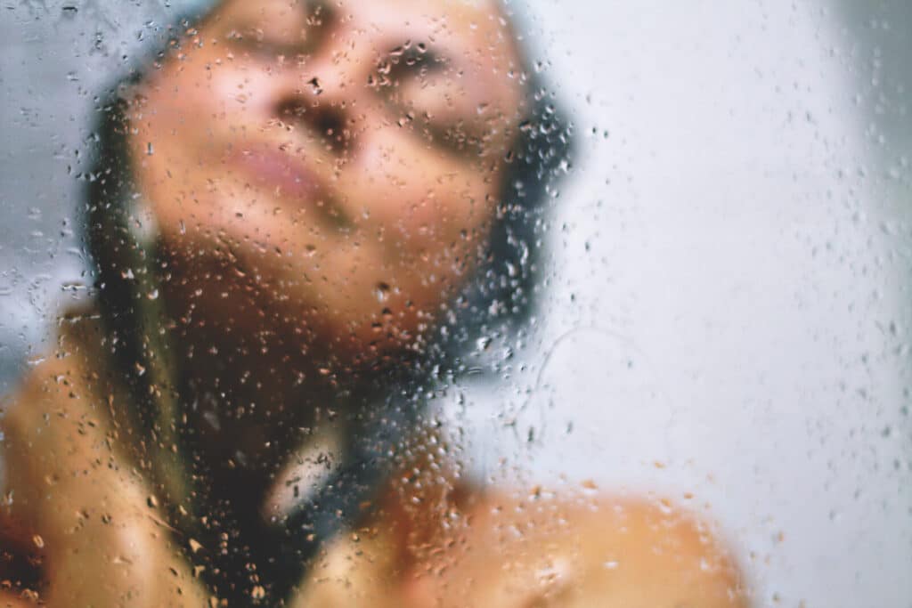 A blurred person stands behind a wet, misty glass surface with water droplets, eyes closed and face relaxed, suggesting they are having a shower or enjoying a moment of calm.