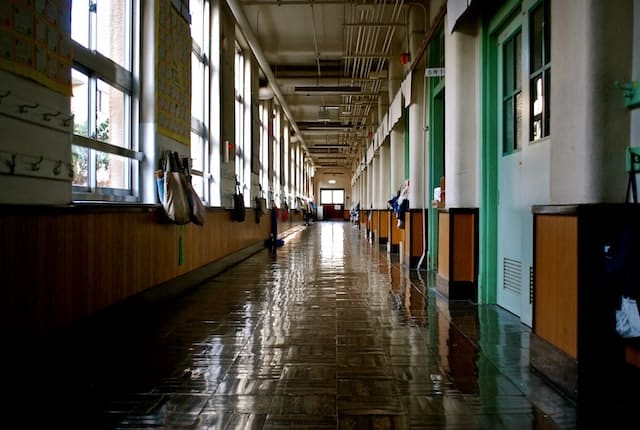 Long, empty school corridor with shiny tiled floor, large windows on the left letting in light, green classroom doors on the right—perhaps leading to a Spanish class—and bags hanging from hooks on the walls.