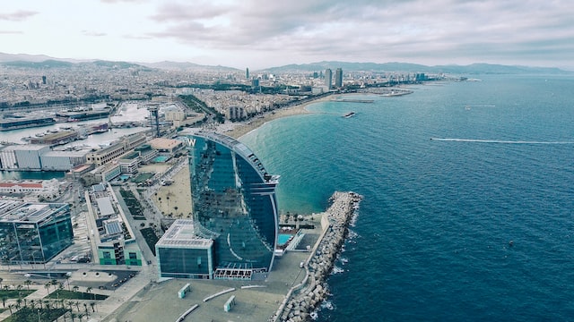 Aerial view of a modern, sail-shaped hotel on the Barcelona coastline, perfect for learning Spanish by the sandy beach and Mediterranean Sea, with the cityscape and mountains in the background under a cloudy sky.