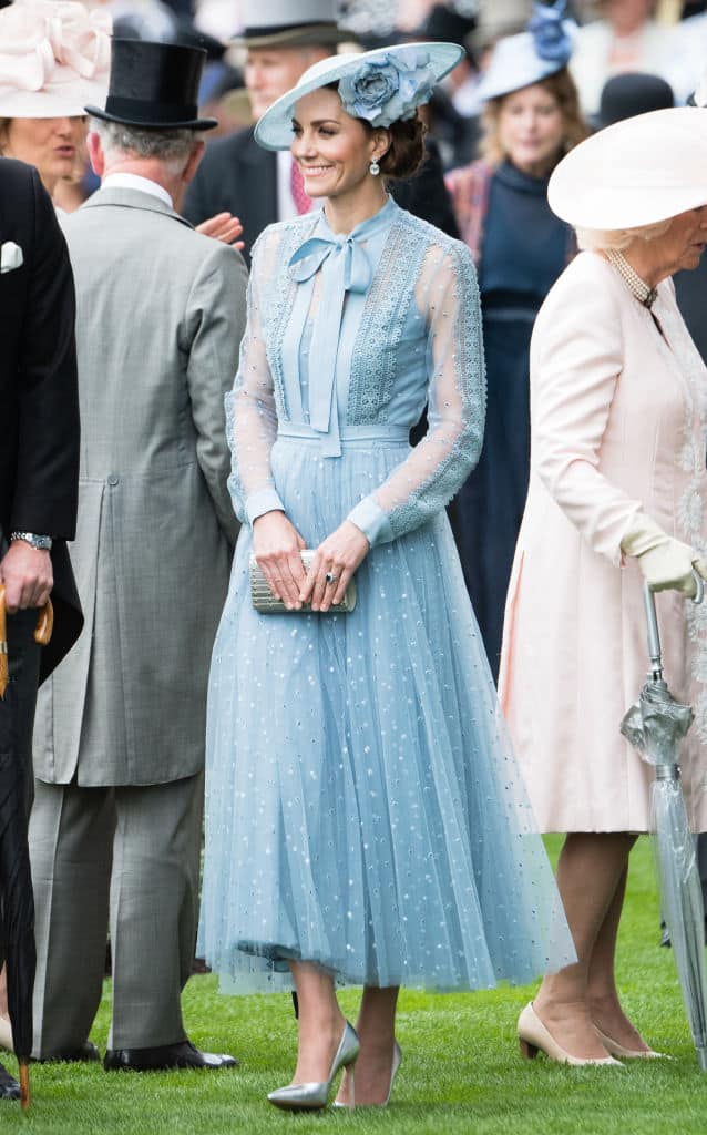 A woman in a light blue, long-sleeved, midi dress with a bow at the neck and a matching wide-brimmed hat stands on grass among others in formal attire at the Cazoo St Leger Festival outdoor event.