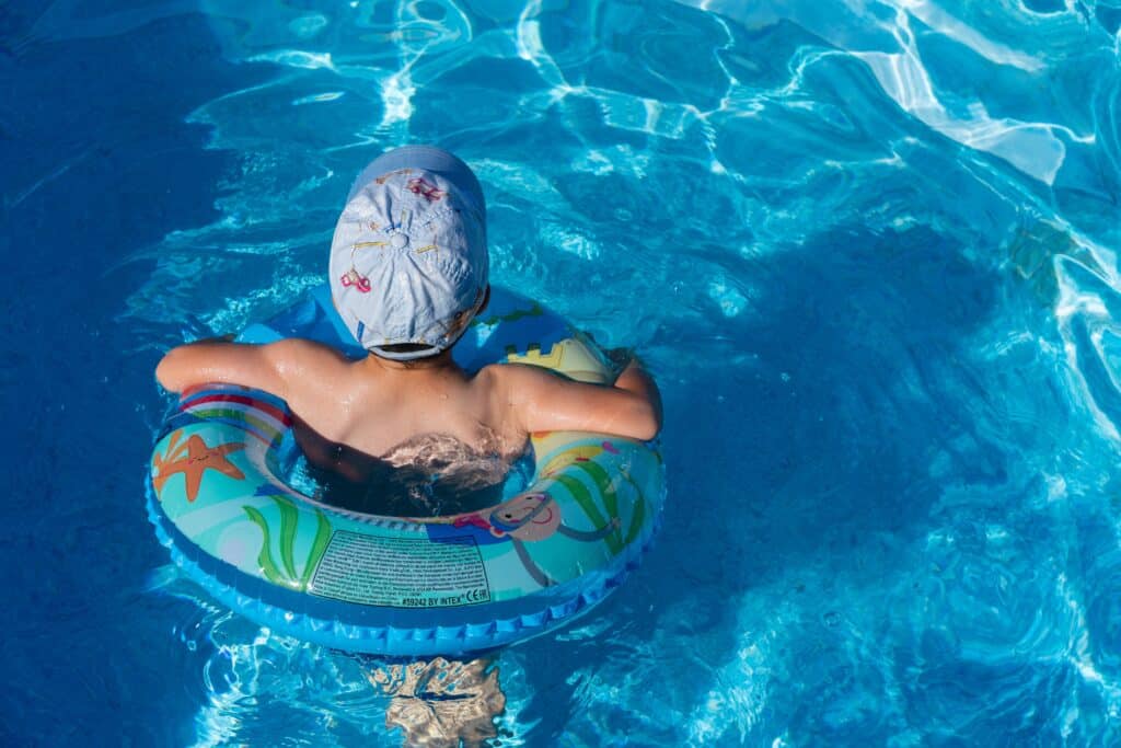 A young child wearing a swimming cap floats in a clear blue pool using an inflatable swim ring decorated with colourful sea creatures, enjoying the water on a sunny family holiday. The scene captures the joy of carefree summer fun.