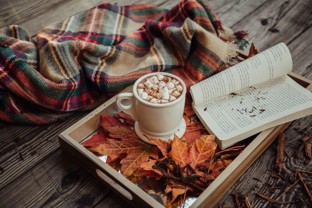 A cup of hot chocolate with marshmallows sits on a tray with autumn leaves, next to an open book and a checked blanket—How To Feel Extra Cosy This Autumn on a rustic wooden surface.
