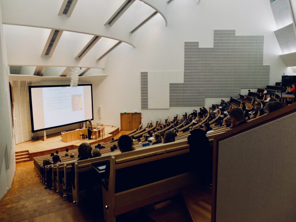 A large university lecture theatre with tiered wooden seating, partially filled with people watching a presentation projected on a screen at the front. The space is modern and well-lit.