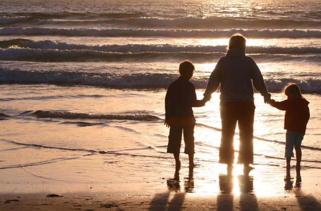 A person and two children hold hands standing on wet sand, facing the sea as the sun sets, with gentle waves and golden light reflecting on the water.