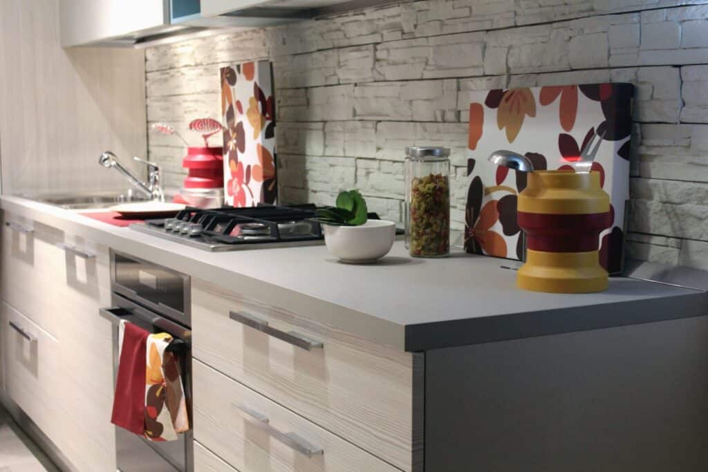 Modern kitchen with light cupboards, a gas cooker, and a sink. Decorative chopping boards with floral patterns, a glass jar with pasta, a white bowl, and a red-yellow folded container sit on the worktop. Stone-patterned splashback.