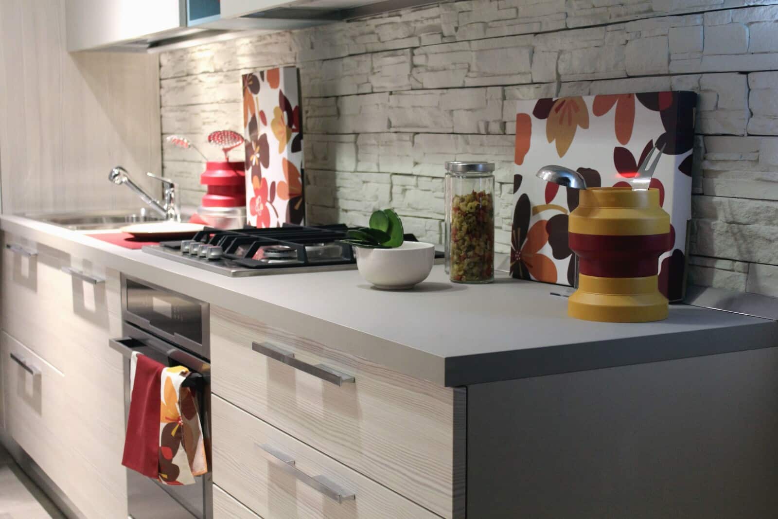 Modern kitchen with light cupboards, a gas cooker, and a sink. Decorative chopping boards with floral patterns, a glass jar with pasta, a white bowl, and a red-yellow folded container sit on the worktop. Stone-patterned splashback.