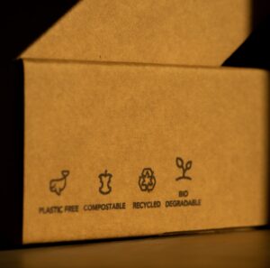 A brown cardboard box displays four eco-friendly icons with labels: "Plastic Free," "Compostable," "Recycled," and "Biodegradable," showing why choose a sustainable packaging company for your business.
