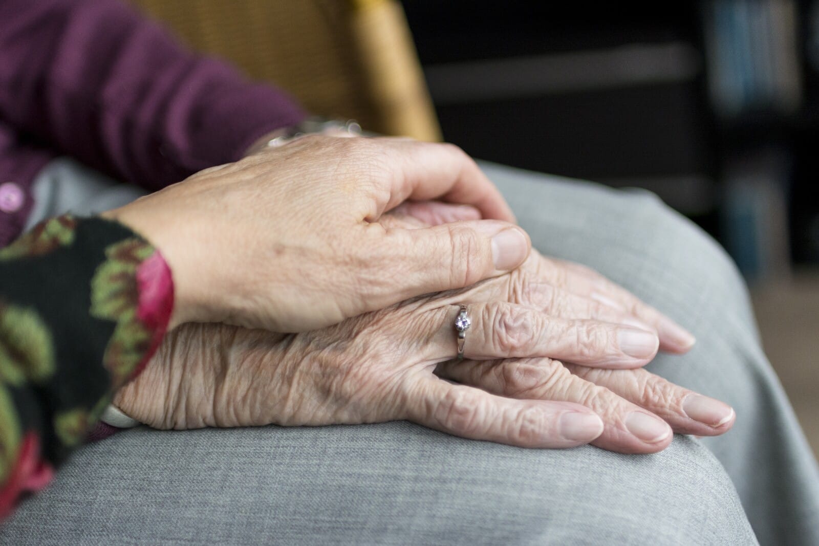 A close-up of one person's hand gently resting on top of an elderly person's hands, who is wearing a ring. Both are seated, creating a comforting and supportive scene.