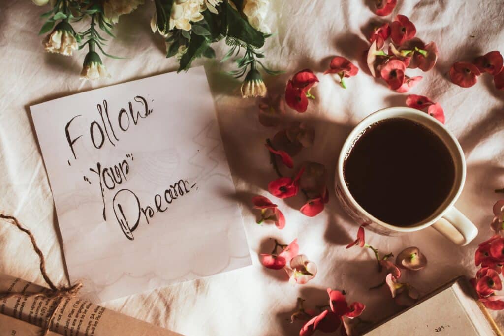 A cup of coffee sits on a white cloth amid red petals, a note reading “Follow Your Dream”, and some white flowers. An open book hints at inspiration—perfect for those wondering how to fulfil a long-term dream as a couple.