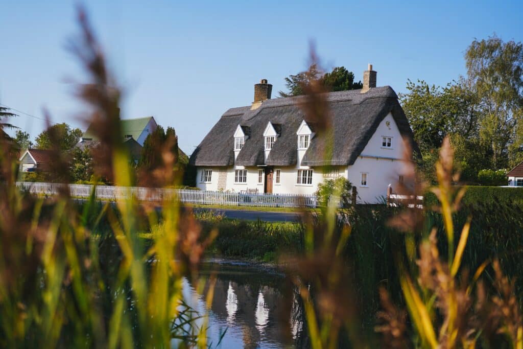 A white cottage with a thatched roof and dormer windows sits behind a pond, surrounded by lush greenery—an inviting scene for anyone seeking inspiration on how to improve the kerb appeal of your house under a clear blue sky.