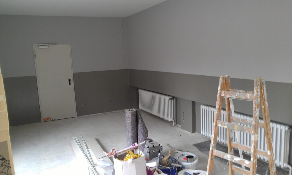 A room under renovation with light grey walls, an unfinished floor, painting supplies and tools scattered around, a wooden ladder, radiators along the wall, and a closed door in the background.