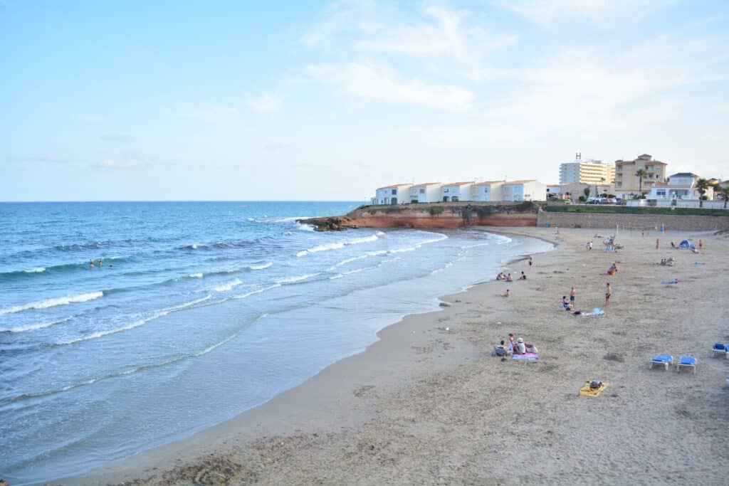 A sandy beach with gentle waves, scattered people sunbathing and swimming, and white buildings along the shoreline—an ideal spot for a relaxing family holiday under a blue sky with light clouds.