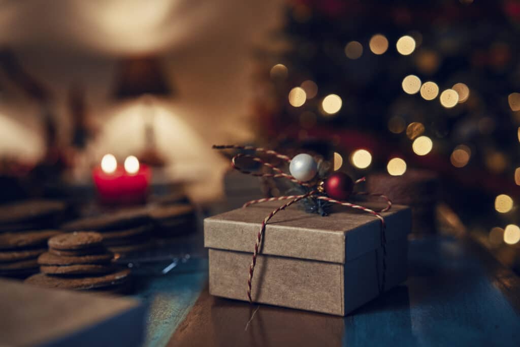 A small, brown gift box tied with string and decorated with festive decorations sits on a table with biscuits and red candles, capturing the cosy spirit of a woman&home Christmas against a backdrop of blurred Christmas lights.
