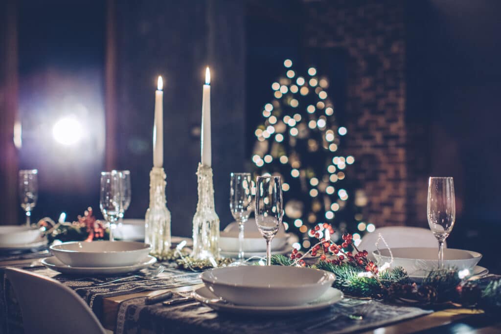 A festive woman&home Christmas dining table set with white plates, wine glasses, two lit candles, and holiday decorations, with a blurred Christmas tree glowing in the background.