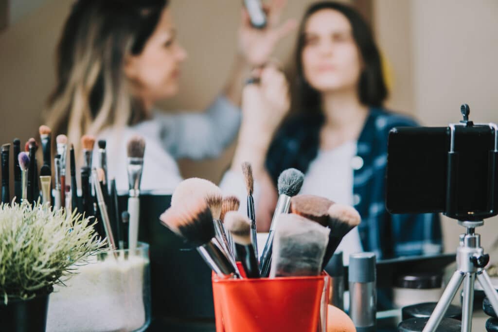 A make-up artist applies festive make-up to a seated woman, perfect for a woman&home Christmas look. In the foreground, brushes and tools sit in containers beside a mobile on a tripod and a small plant. The softly blurred background adds warmth.