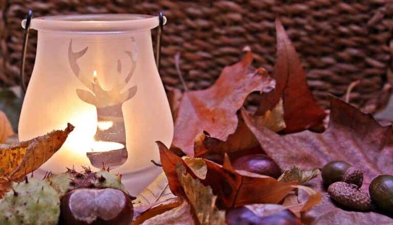 A lit candle in a frosted glass holder with a reindeer design sits among autumn leaves, conkers, and acorns, with a wicker basket in the background.