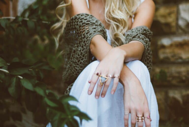 A woman with long blonde hair sits outdoors, wearing a white dress and a green knitted shawl. She ponders what to do when jewellery gets damaged, her multiple rings sparkling as she crosses her arms over her knees by a stone wall with green plants behind her.