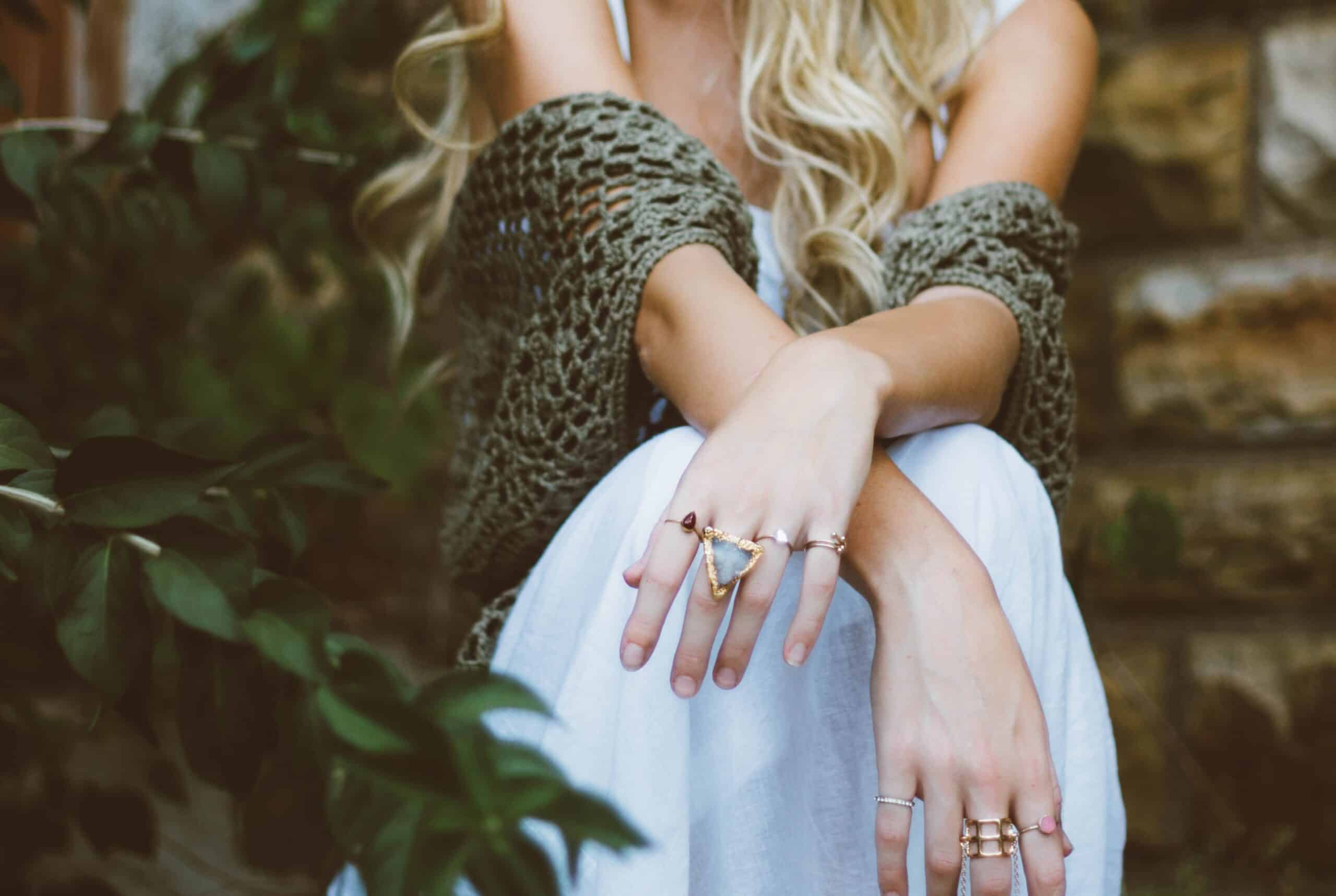 A woman with long blonde hair sits outdoors, wearing a white dress and a green knitted shawl. She ponders what to do when jewellery gets damaged, her multiple rings sparkling as she crosses her arms over her knees by a stone wall with green plants behind her.