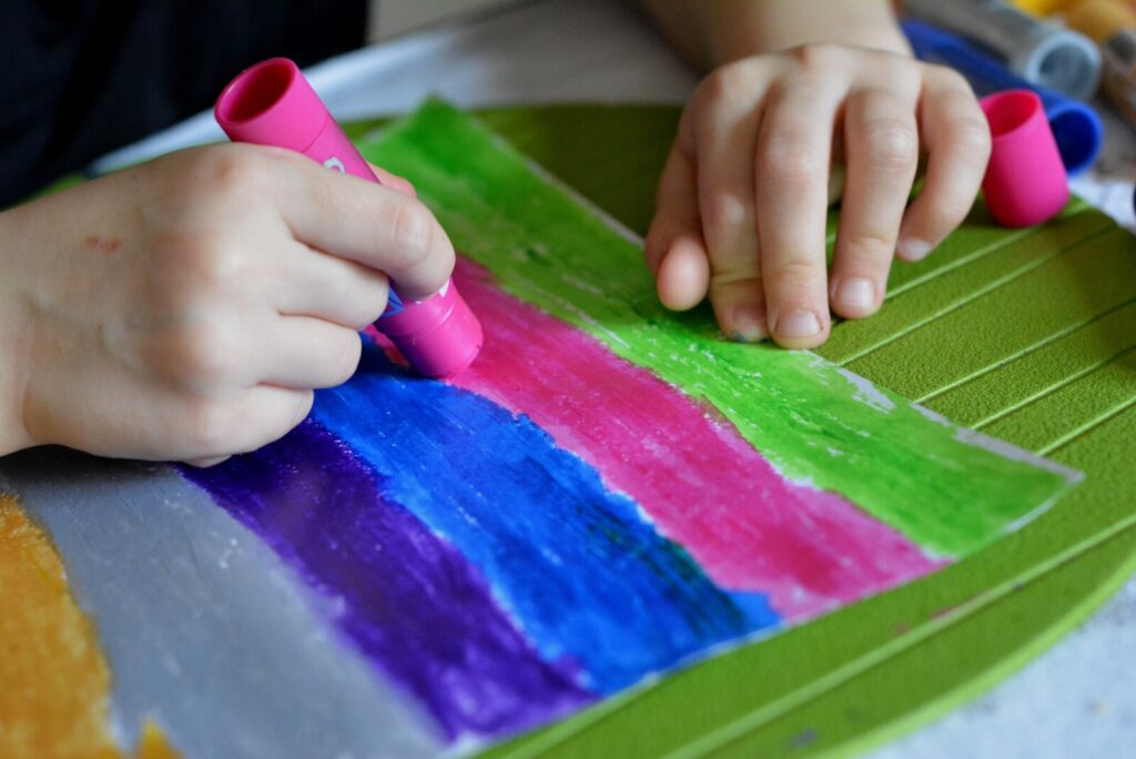 A child’s hands colouring a sheet of paper with wide, colourful stripes using thick crayons; green, blue, pink, and purple colours are visible. The paper is placed on a green surface.