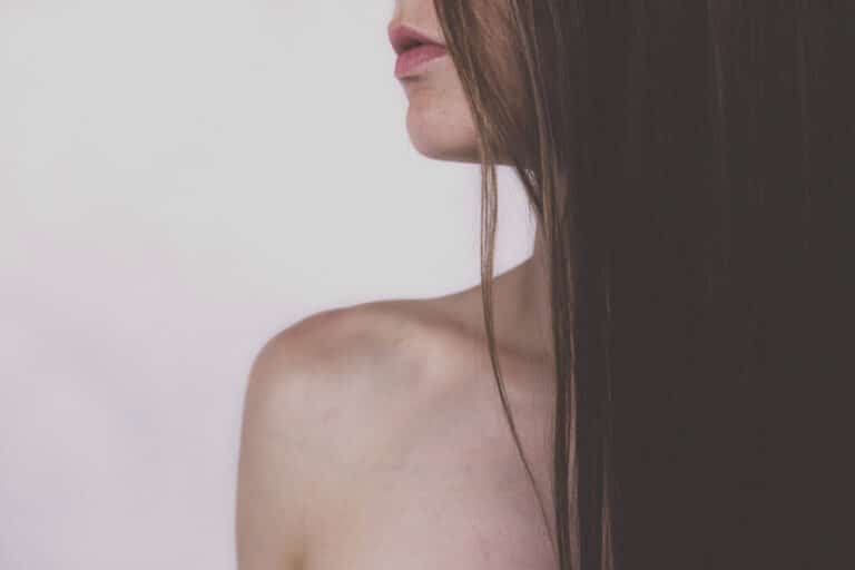 A close-up of a woman's bare shoulder and neck, with long, straight brown hair partially covering her chest and face, set against a plain light background. Her lips are slightly parted.
