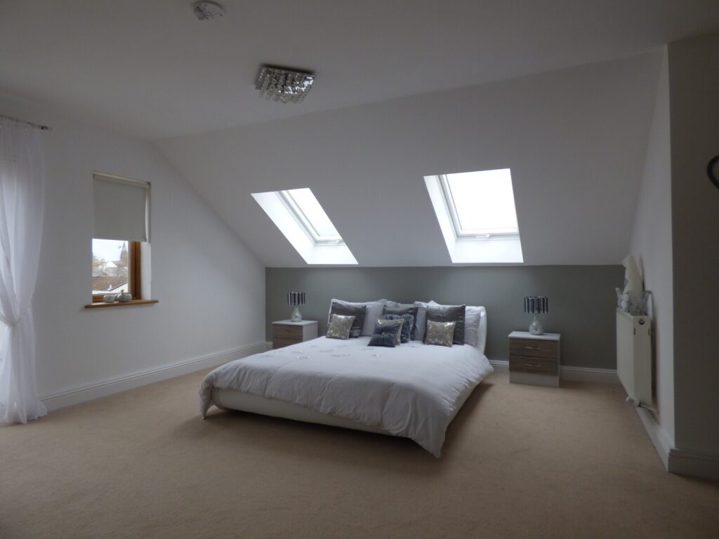 A modern loft bedroom with a low double bed, white bedding, two bedside tables, and rooflight windows letting in natural light. The walls are white with a muted green accent behind the bed.