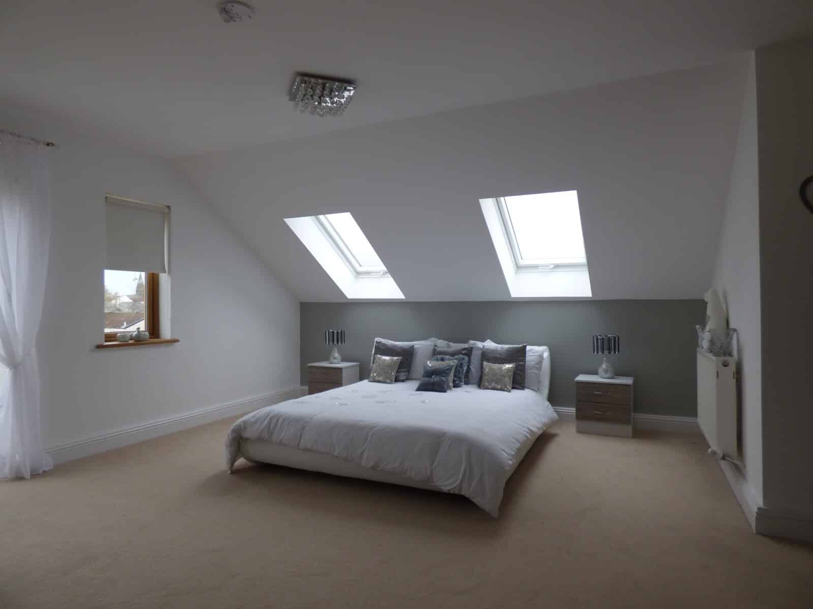 A modern loft bedroom with a low double bed, white bedding, two bedside tables, and rooflight windows letting in natural light. The walls are white with a muted green accent behind the bed.
