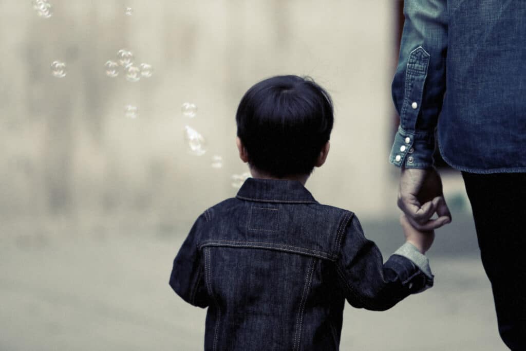 A young child in a denim jacket holds an adult's hand whilst walking. The adult also wears denim. Bubbles float nearby against a blurred background, creating a gentle, peaceful scene.