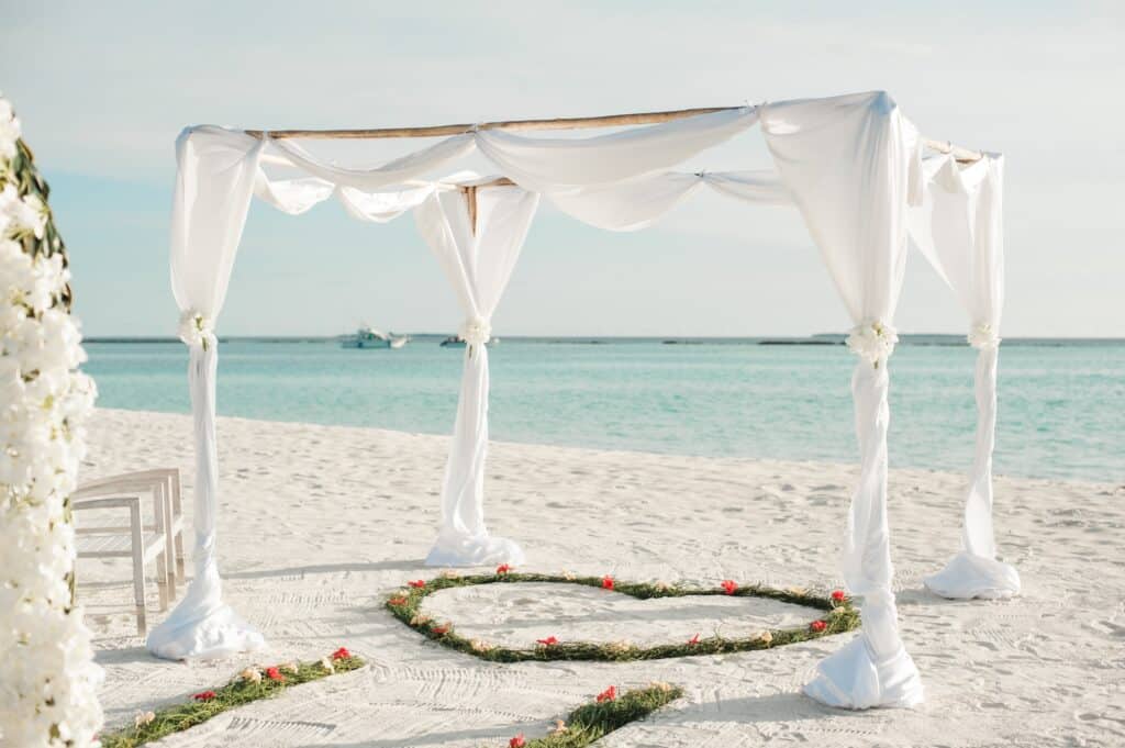 A white wedding canopy with flowing drapes stands on a sandy beach near the sea, showing How To Plan the Perfect Beach Wedding, with a heart-shaped decoration of greenery and red flowers on the sand beneath it.