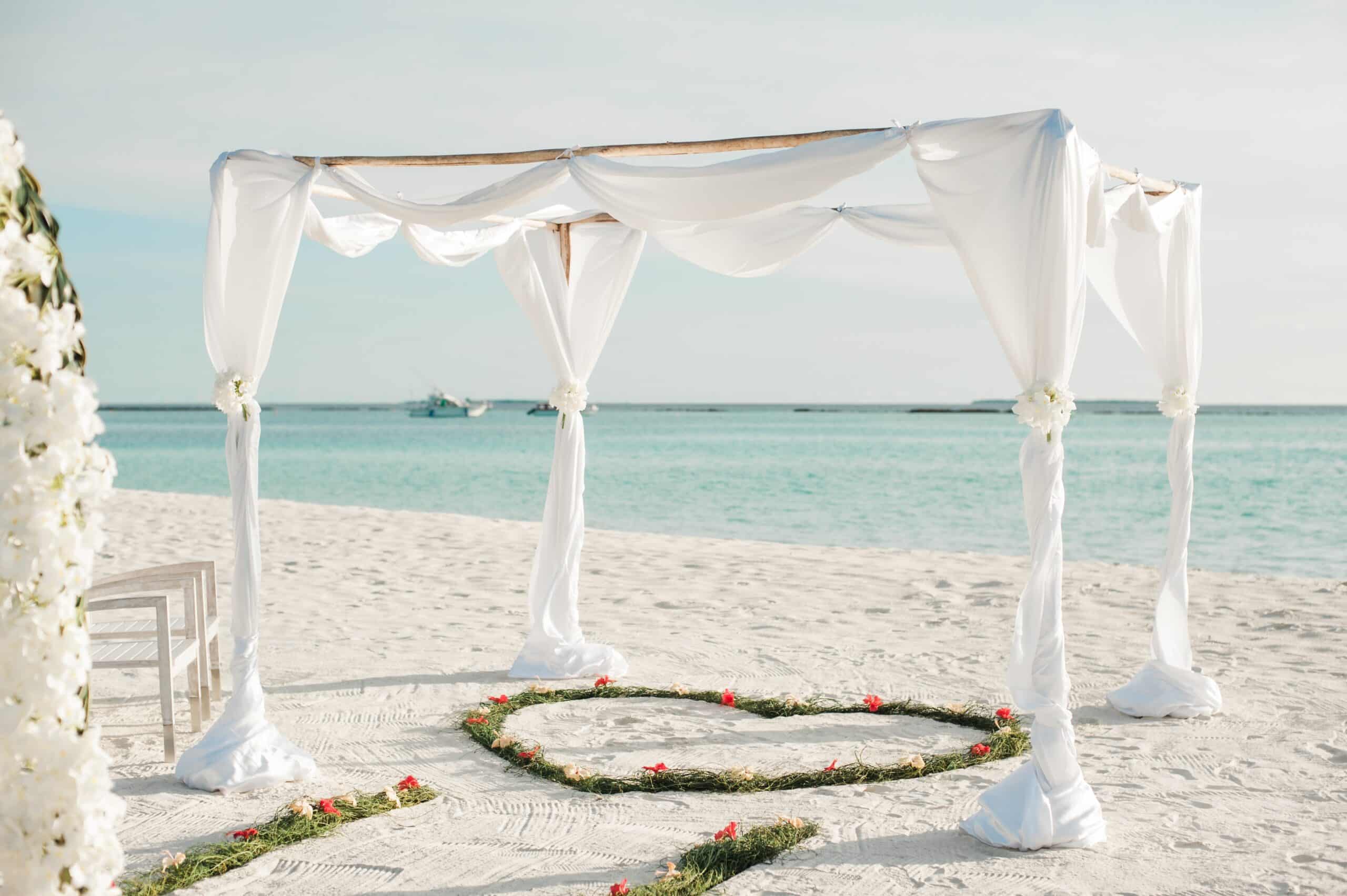 A white wedding canopy with flowing drapes stands on a sandy beach near the sea, showing How To Plan the Perfect Beach Wedding, with a heart-shaped decoration of greenery and red flowers on the sand beneath it.