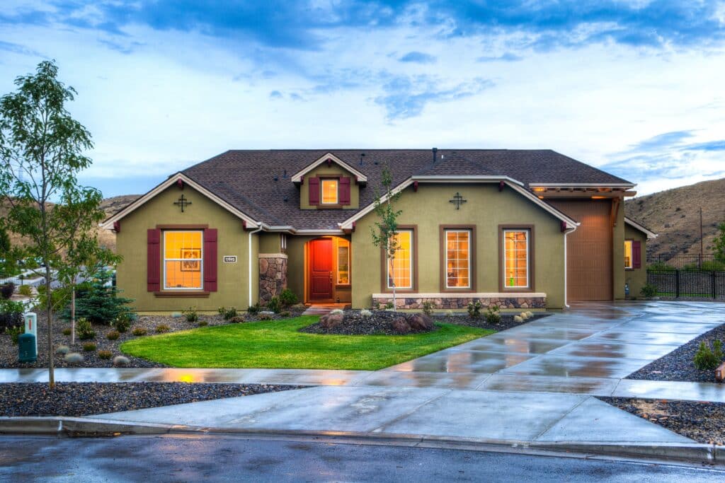 Single-storey house with green exterior, brown shutters, and warmly lit windows. A wet driveway and pavement reflect light—showcasing 4 Top Tips to Update Your Kerb Appeal for an Aesthetically Pleasing Home amid hills under a cloudy sky.