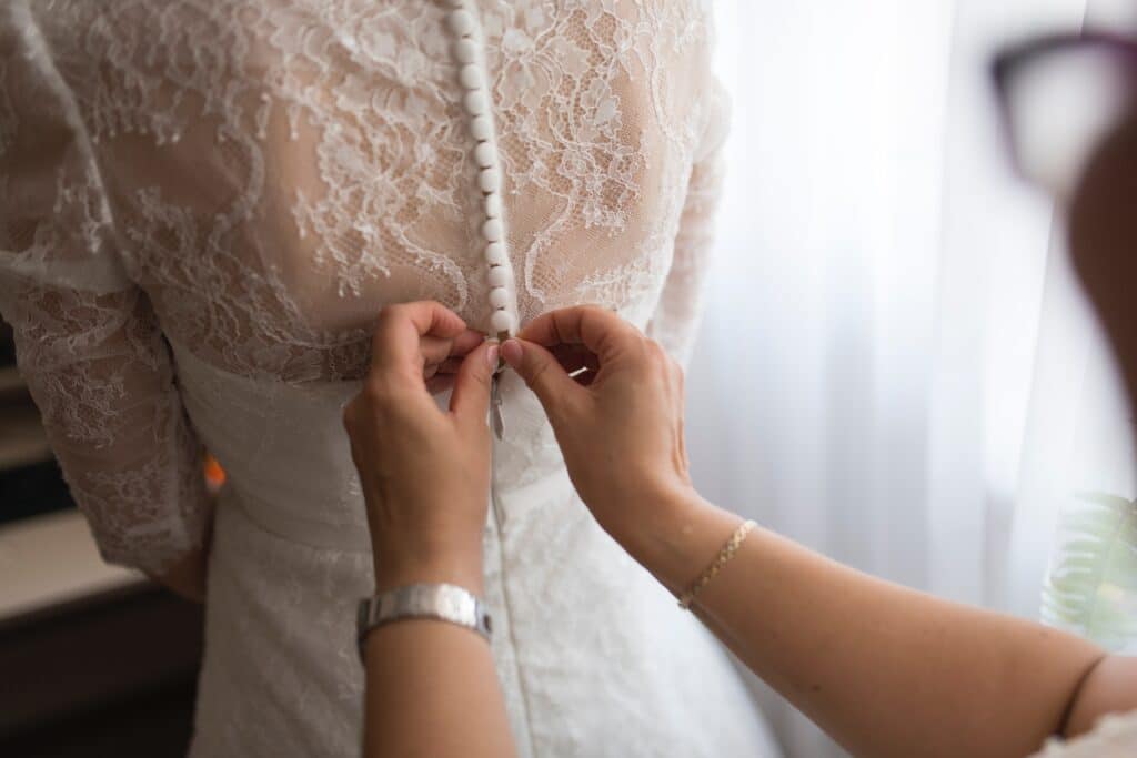 A person fastens white buttons on the back of a bride’s lace wedding dress, focussing on intricate lace detail and delicate hands—an intimate moment that may evoke thoughts like, How Much Influence Should A Parent Have Over Their Child's Marriage?.