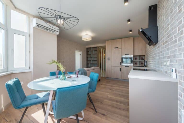 Modern open-plan kitchen and dining area with light wood laminate floors—showing why laminate flooring serves as a good basis for a refurbishment—blue chairs around a round table, large windows, built-in cupboards, and exposed brick wall. Bright and contemporary space.