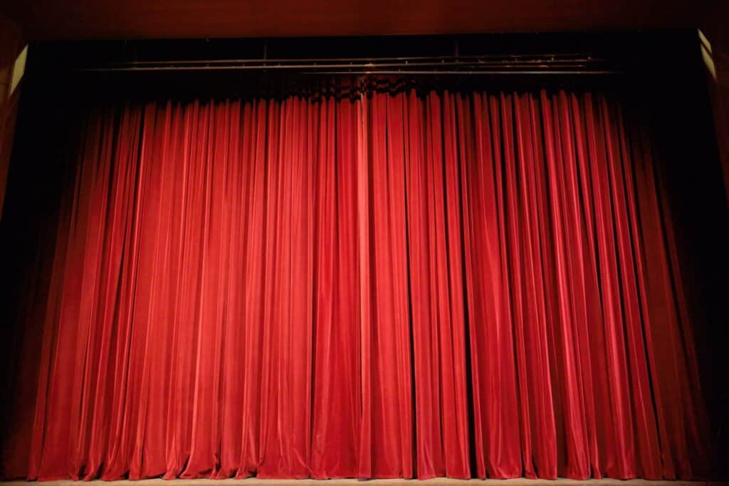 A large stage with closed, deep red velvet curtains, evenly pleated from top to bottom, under dim theatre lighting.